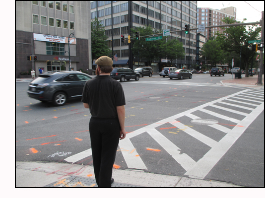 Photo shows a man standing on a corner facing a 4-lane street.  To his left is a 6-lane street with a vehicle half-way through the intersection, going the same direction he's facing.
